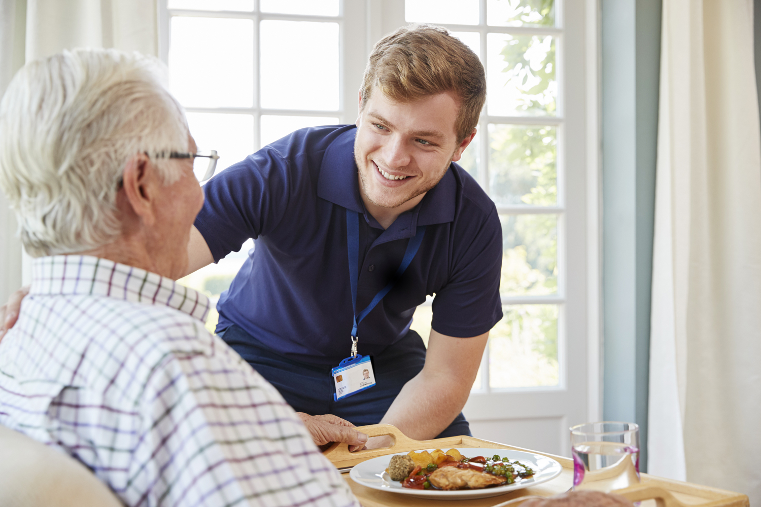Bilden föreställer en yngre man i blå vårdkläder som vänskapligt håller handen på en äldre mans axel efter att precis ha serverat honom en bricka med mat.