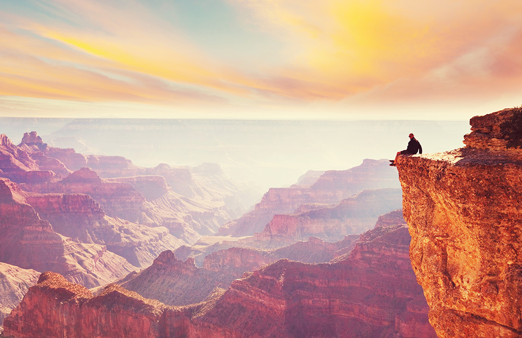 Människa sitter på en klippa och tittar ut över ett vackert, soligt berglandskap